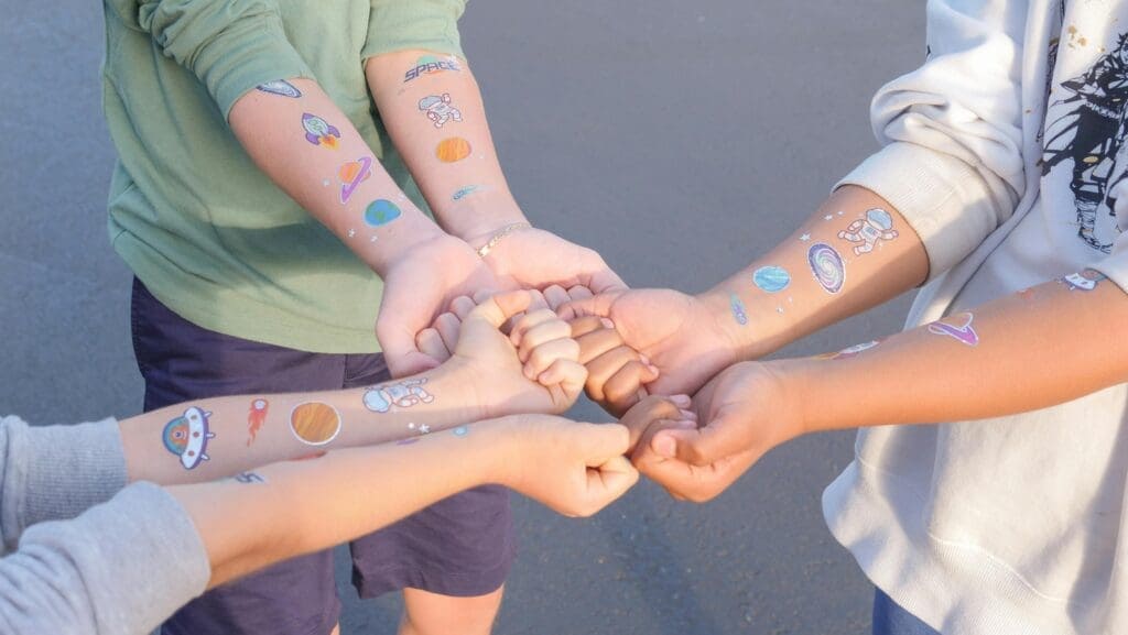 Das Bild zeigt 3 Kinder auf dem Schulhof, die sich mit ausgetreckten Armen gegenüber stehen. Für uns symbolisiert das eine demokratische Abstimmung/Einigung.