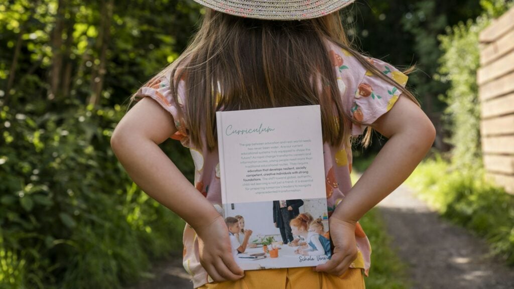 Warum unsere Schule keine Noten, Fächer oder Jahrgänge braucht (Newsletter 5/2025) 3 Ein Kind, dass das Curriculum von Schola Vera in Buchform hinter ihrem Rücken hält.